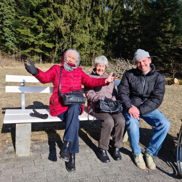Zwei ältere Frauen und Rollstuhl, lachend und junger Mensch auf Sitzbank in der Sonne